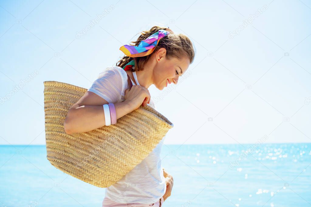 happy modern 40 year old woman in white t-shirt with beach straw bag on the beach.
