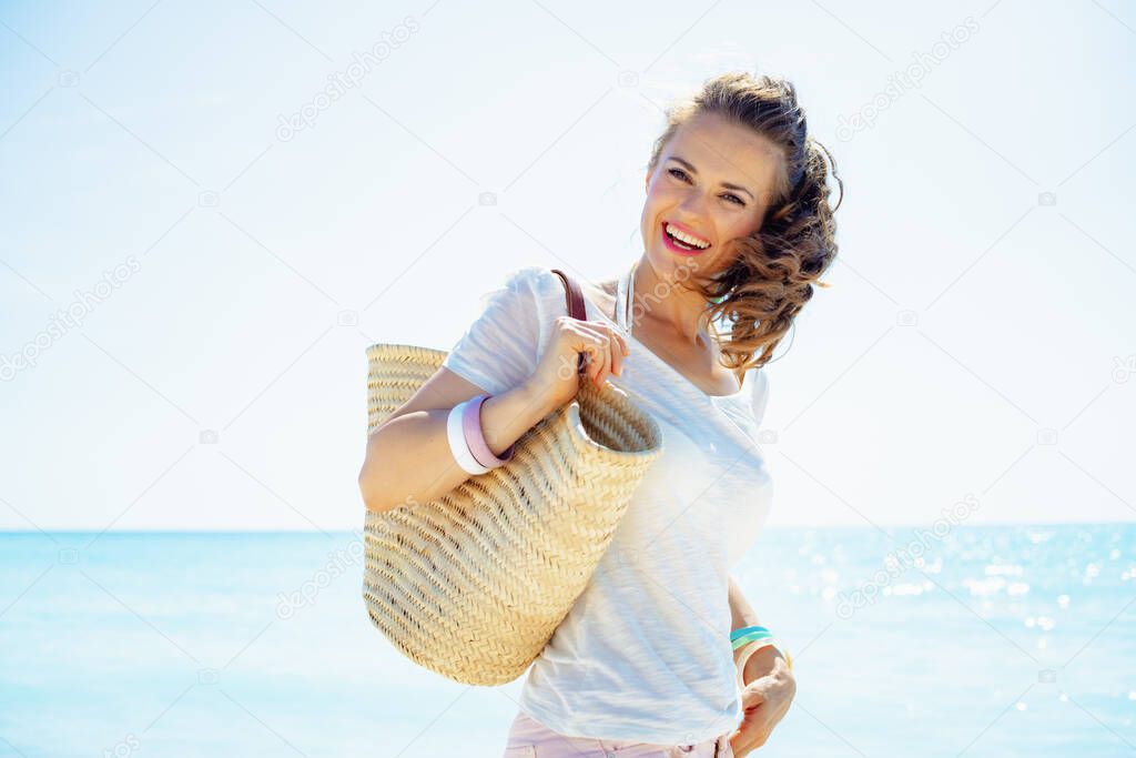 Portrait of smiling young woman in white t-shirt with beach straw bag on the beach.