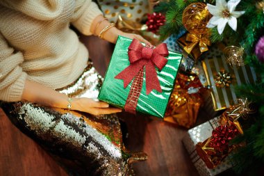 Altın pullu etekli ve beyaz süveterli genç bayana yakın çekim. Noel hediyesi kutusunun yanındaki süslenmiş Noel ağacının altında..