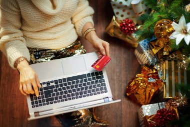 Altın pullu etekli, beyaz kazaklı, elektronik alışveriş sitesinden online alışveriş yapan ve hediye kutularının yanında süslenmiş Noel ağacının altında şık bir ev hanımı..
