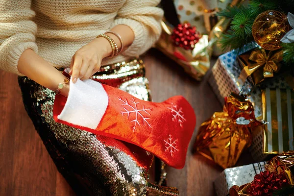 身着金色紧身衣裙和白色毛衣的年轻女子在装饰圣诞树下的衣服 就在礼物盒旁边 从红色圣诞长袜里拿出一些东西 — 图库照片