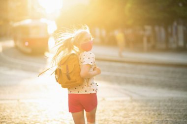 Koronavirüs salgını sırasında hayat. Beyaz benekli bluzlu, pembe maskeli ve sarı sırt çantalı modern bir öğrenci okuldan dönüyor..