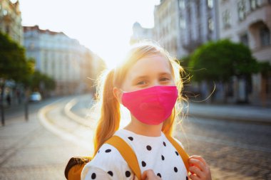 Koronavirüs salgını sırasında hayat. Beyaz benekli bluzlu, pembe maskeli ve sarı sırt çantalı modern öğrencinin portresi dışarıda okula gidiyor..