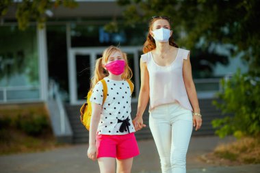 Koronavirüs salgını sırasında hayat. Maskeli ve sarı sırt çantalı zarif anne ve çocuk dışarıda okula gidiyor..