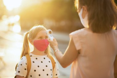 Maskeli modern bayan öğretmen ve öğrenci dışarıdaki dijital termometreyle sıcaklığı ölçüyorlar..