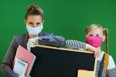 Maskeli ve ders kitabı olan modern bayan öğretmen ve öğrenci kara tahtada yeşil arka planda izole edilmiş boş kara tahtayı gösteriyor..