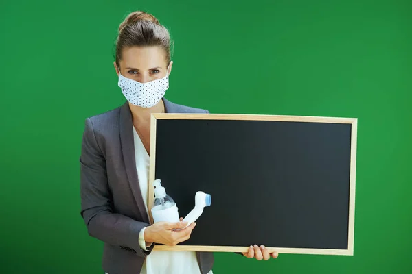 Covid 19大流行病期间的生活 身穿白衬衫的现代女教师 戴着医疗面罩 带数字温度计和抗菌剂 在黑板绿色背景下显示空白黑板 — 图库照片