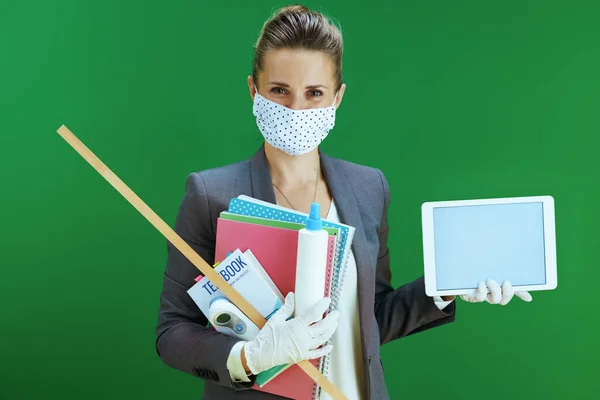 modern female teacher in white blouse with medical mask, rubber gloves, digital thermometer, textbook and sanitizer showing tablet PC blank screen against chalkboard green background.