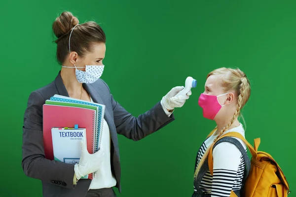 modern female teacher and pupil with masks, yellow backpack and textbook measures temperature with digital thermometer isolated on chalkboard green.