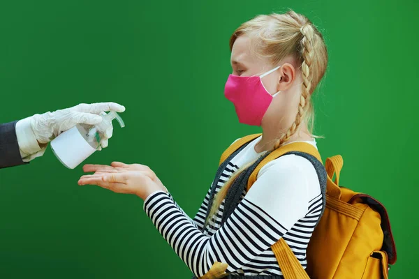 modern female teacher and pupil with mask and yellow backpack disinfecting hands with sanitizer on chalkboard green background.