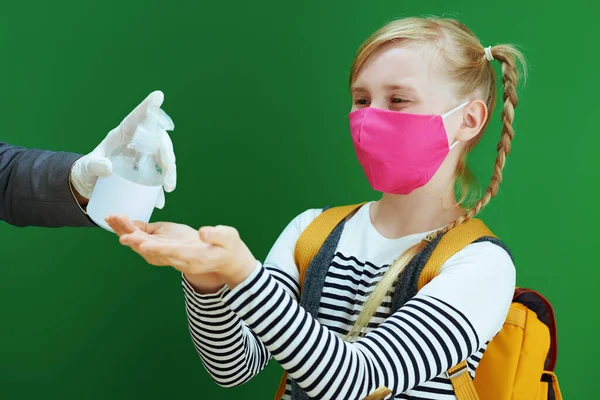 modern female teacher and pupil with mask and yellow backpack disinfecting hands with sanitizer isolated on chalkboard green background.