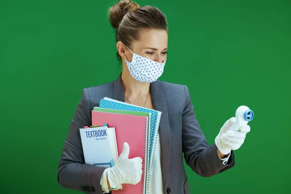 modern woman pedagogue in white blouse with medical mask, rubber gloves and textbook measures temperature with digital thermometer showing thumbs up isolated on chalkboard green.