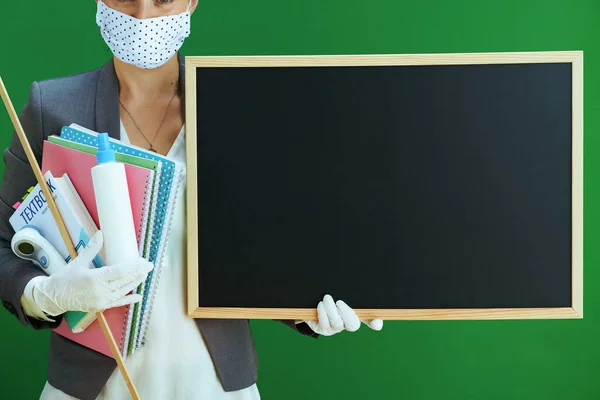 Closeup on female teacher in white blouse with medical mask, rubber gloves, digital thermometer, textbook and an antibacterial agent showing blank blackboard isolated on chalkboard green.