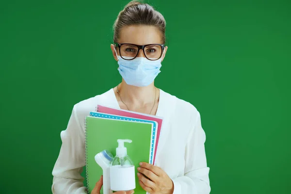 Life during covid-19 pandemic. Portrait of modern woman pedagogue in white blouse with medical mask, digital thermometer and sanitizer against chalkboard green background.