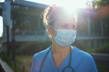 Koronavirüs salgını. Steteskop ve tıbbi maskeli, önlüklü, üzgün, modern hekim kadın kliniğin dışında..