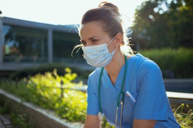 Koronavirüs salgını. Steteskop ve tıbbi maskeli, üniformalı, düşünceli, modern hekim kadın kliniğin yakınında oturuyor..