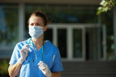 Koronavirüs salgını. Steteskop ve tıbbi maske takmış kendine güvenen modern doktor kadın hastanenin yakınındaki açık havaya bakıyor..