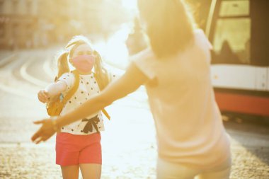 Koronavirüs salgını sırasında hayat. Gülümseyen zarif anne ve çocuk maskeli ve sarı sırt çantalı okuldan dışarı çıkıyor..