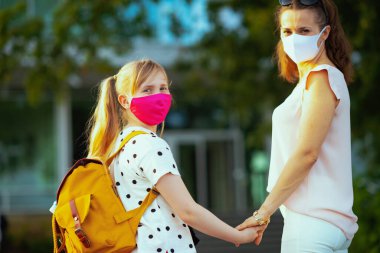 Koronavirüs salgını sırasında hayat. Açık havada okula giden maskeli ve sarı sırt çantalı genç anne ve okul öğrencisinin portresi..