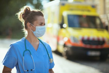 covid-19 pandemic. pensive modern medical doctor woman in uniform with stethoscope and medical mask outside near ambulance. clipart