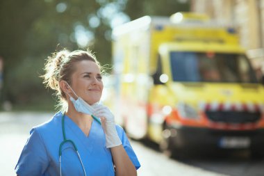 Koronavirüs salgını. Steteskoplu ve tıbbi maskeli modern doktor kadın dışarıda ambulansın yanında nefes alıyor..