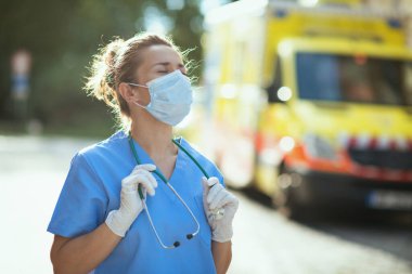Koronavirüs salgını. Dışarıda ambulansın yanında steteskop ve maske takan yorgun, modern sağlık görevlisi kadın..