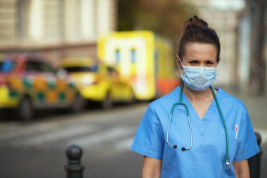 coronavirus pandemic. Portrait of modern paramedic woman in uniform with stethoscope and medical mask outside near ambulance. clipart