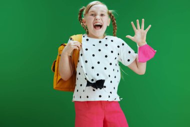 Koronavirüs salgını sırasında hayat. Sırt çantalı, pembe maskeli, beyaz benekli bluzlu, atlayan ve el sallayan, tebeşir yeşili arka planda yalnız gülümseyen modern kız..