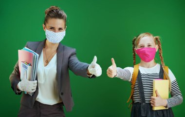 Gülümseyen modern öğretmen kadın ve maskeli okul kızı kara tahta yeşil arka planda başparmak gösteriyorlar..