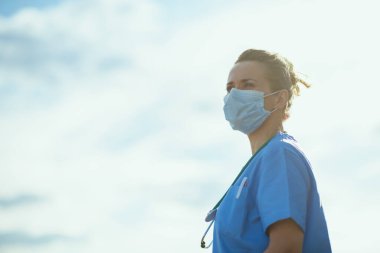 Koronavirüs salgını. Kendine güvenen, doktor önlüklü, tıbbi maskeli bir kadın şehrin dışında gökyüzüne karşı mesafeye bakıyor..