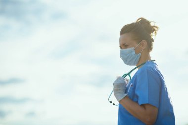 Koronavirüs salgını. Steteskoplu, tıbbi maskeli ve dışarıda gökyüzüne karşı lastik eldivenli, düşünceli, modern tıbbi pratisyen kadın..