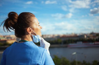 Covid-19 salgını. Şehirde gökyüzüne karşı nefes alan tıbbi maskeli ve lastik eldivenli rahat modern hekim kadın..