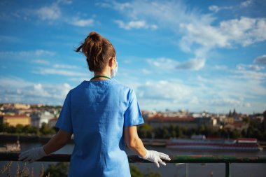 Koronavirüs salgını. Tıbbi maskeli ve lastik eldivenli modern tıp pratisyeni kadın gökyüzüne doğru dışarı bakıyor..