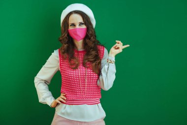 Koronavirüs salgını sırasında hayat. Pembe kolsuz gömlekli, pembe tıbbi maskeli zarif kadın yeşil arka planda izole edilmiş bir şeyi işaret ediyor..