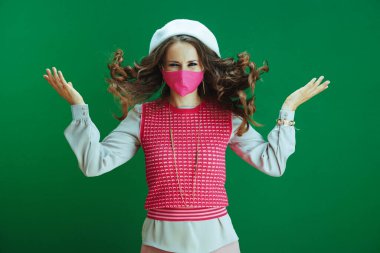 Koronavirüs salgını sırasında hayat. Pembe maskeli, beyaz bereli, yeşil arka planda zıplayan gülümseyen genç bayan öğrenci..