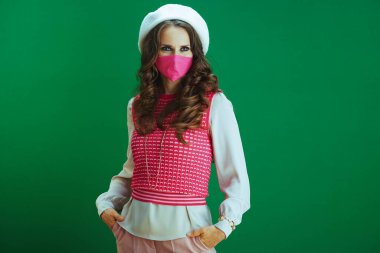 Koronavirüs salgını sırasında hayat. Pembe kolsuz gömlekli, yeşil arka planda pembe tıbbi maskeli şık bir öğrenci portresi..