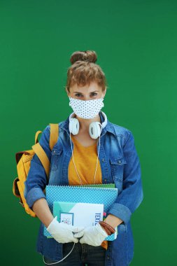 Koronavirüs salgını sırasında hayat. Ders kitapları, defterler, tıbbi maske, beyaz kulaklıklar ve yeşil arka planlı sırt çantalı modern öğrenci..