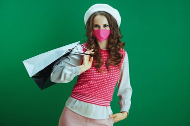 Koronavirüs salgını sırasında hayat. Pembe kolsuz gömlekli, yeşil arka planda pembe tıbbi maskeli ve alışveriş çantalı zarif kadın portresi..