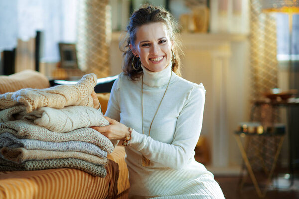 smiling stylish female in white sweater and skirt sitting near couch with pile of sweaters in the modern house in sunny winter day.