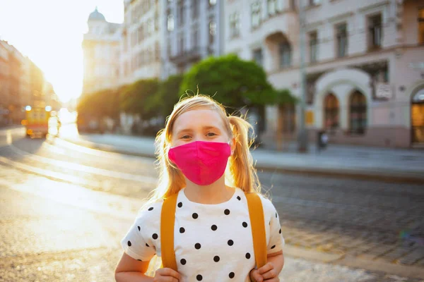 Koronavirüs salgını sırasında hayat. Beyaz benekli bluzlu, pembe maskeli ve sarı sırt çantalı modern bir çocuğun portresi okuldan dönüyordu..