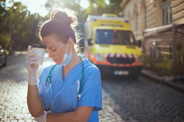covid-19 pandemic. tired modern medical doctor woman in uniform with stethoscope, medical mask and cup of coffee outside near ambulance.