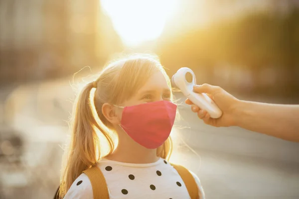modern teacher woman and school girl with mask measures temperature with digital thermometer outdoors.