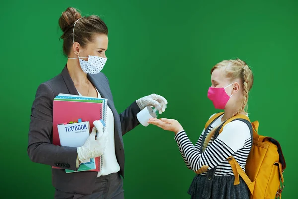带有面具 黄色背包和课本的现代女教师和女学生在黑板绿色背景下用清洁剂消毒手 — 图库照片