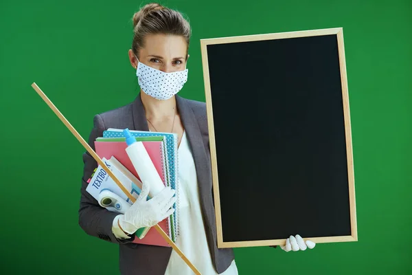 modern woman pedagogue in white blouse with medical mask, rubber gloves, digital thermometer, textbook and an antibacterial agent showing blank board isolated on chalkboard green background.