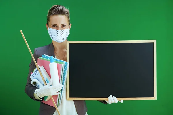 身穿白衬衫的现代女教师 戴着医疗面罩 橡胶手套 数字温度计 课本和卫生纸 展示了在黑板绿色背景上隔离的空白黑板 — 图库照片