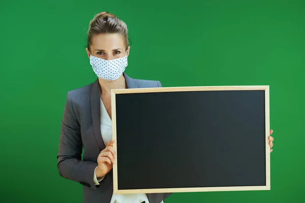 Covid 19大流行病期间的生活 现代女教师 身穿白衬衫 戴着医疗面罩 在黑板绿色背景下展示空白黑板 — 图库照片