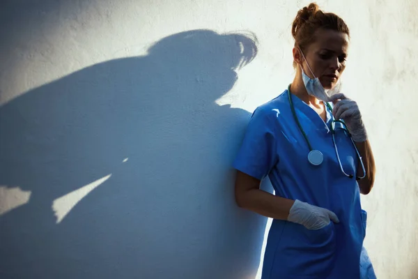 コロナウイルスのパンデミック 壁の近くの街で聴診器 医療用マスク ゴム手袋を持つスクラブの不幸な現代の医師の女性 — ストック写真