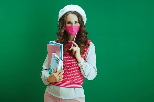 コロナウイルスのパンデミックの間の生活 緑の背景に教科書やノート ピンクの医療用マスクを持つ白いベレー帽の高価なスタイリッシュな学生 — ストック写真