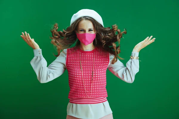 Koronavirüs salgını sırasında hayat. Pembe maskeli, beyaz bereli, yeşil arka planda zıplayan gülümseyen genç bayan öğrenci..