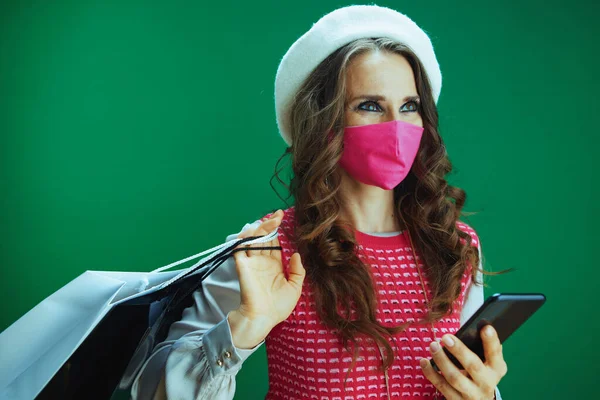 コロナウイルスのパンデミックの間の生活 ピンクの医療用マスクとショッピングバッグを持つ白いベレー帽のスタイリッシュな女性の買い物客は緑の背景に隔離されたスマートフォンでオンラインで購入 — ストック写真